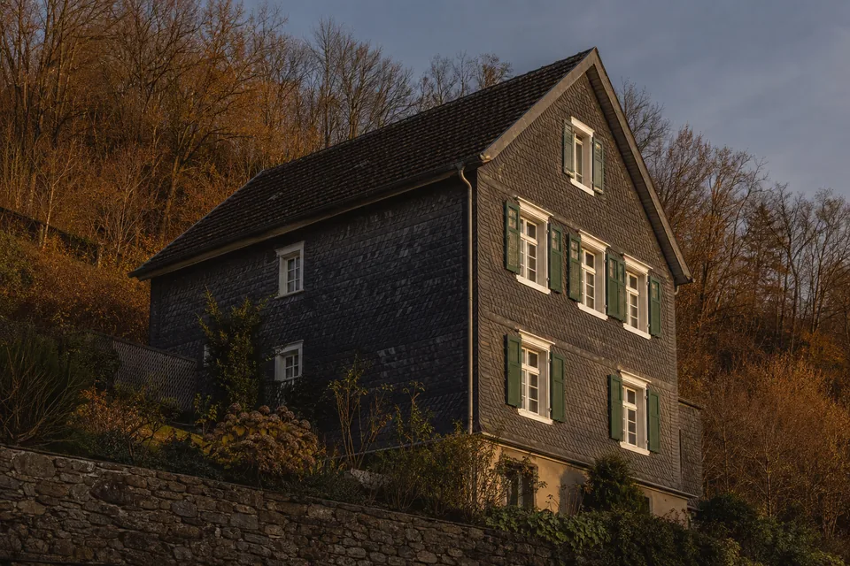 Verschiefertes Wohnhaus am Hang, weiß abgesetzte Fenstergewände und eine Bruchsteinstützmauer im Vordergrund.